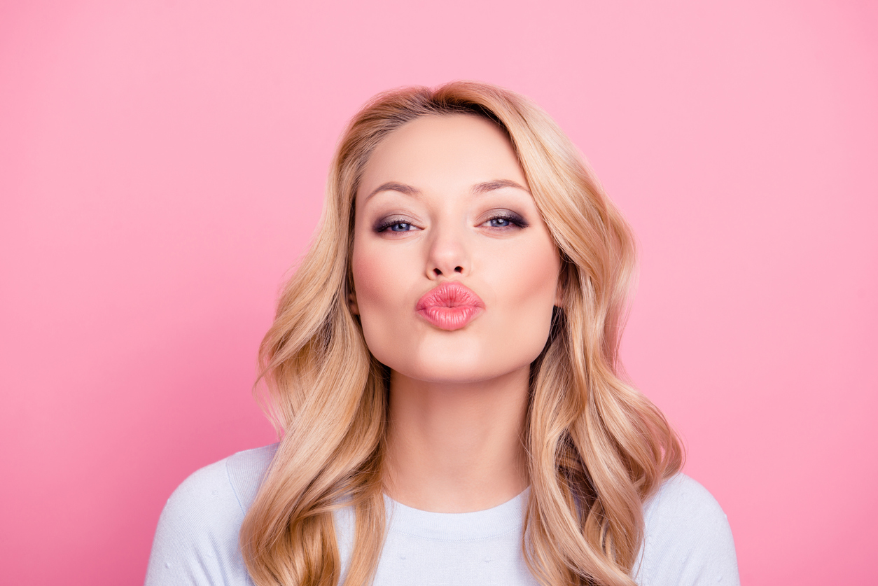 A beautiful blonde woman blowing a kiss against a soft pink background, exuding charm and warmth.