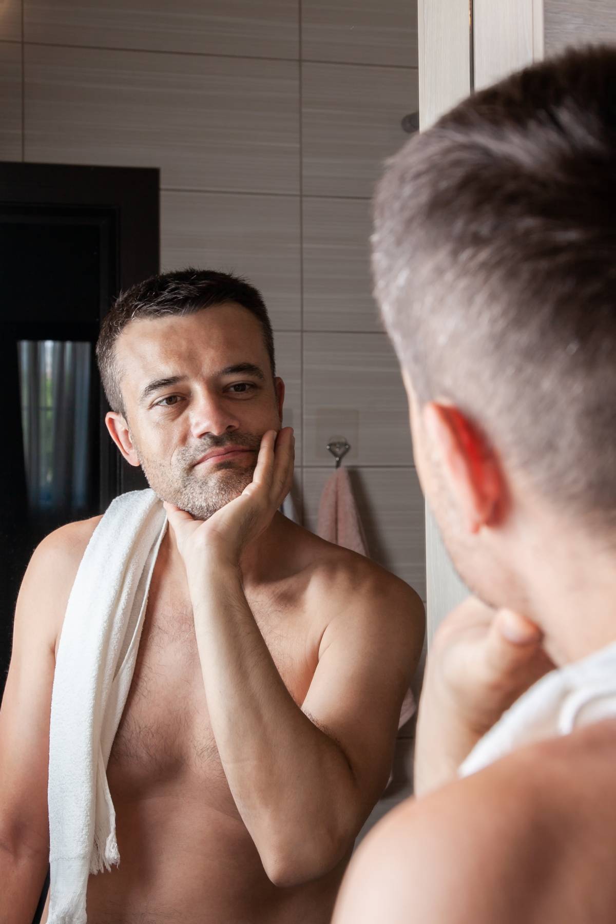A man looks in the mirror and touches his chin with stubble