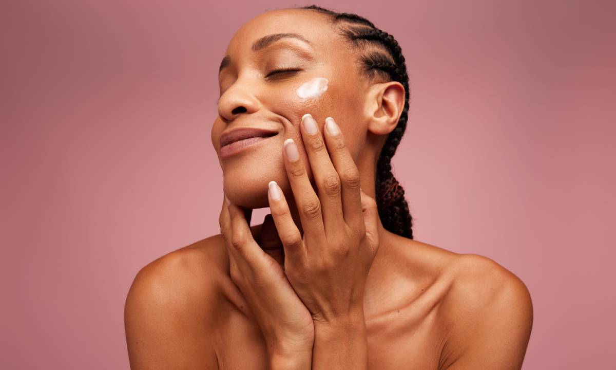 A woman with her hands on her face, set against a soft pink background, conveying a sense of contemplation or surprise.